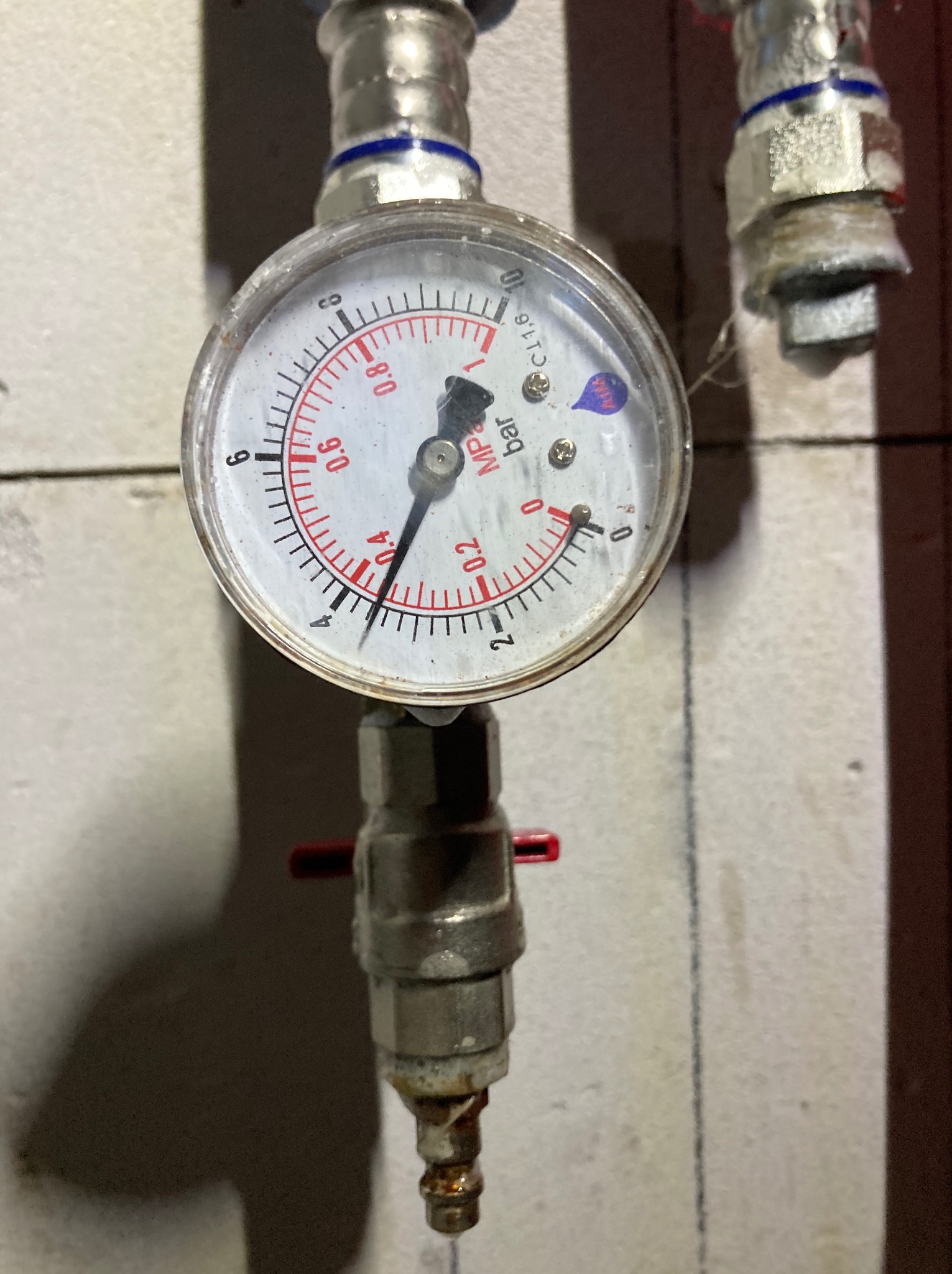 Manometr z zaworem na instalacji, wskazujący ciśnienie w barach. Widoczne złącza i fragment ściany. Detal instalacji hydraulicznej lub grzewczej.