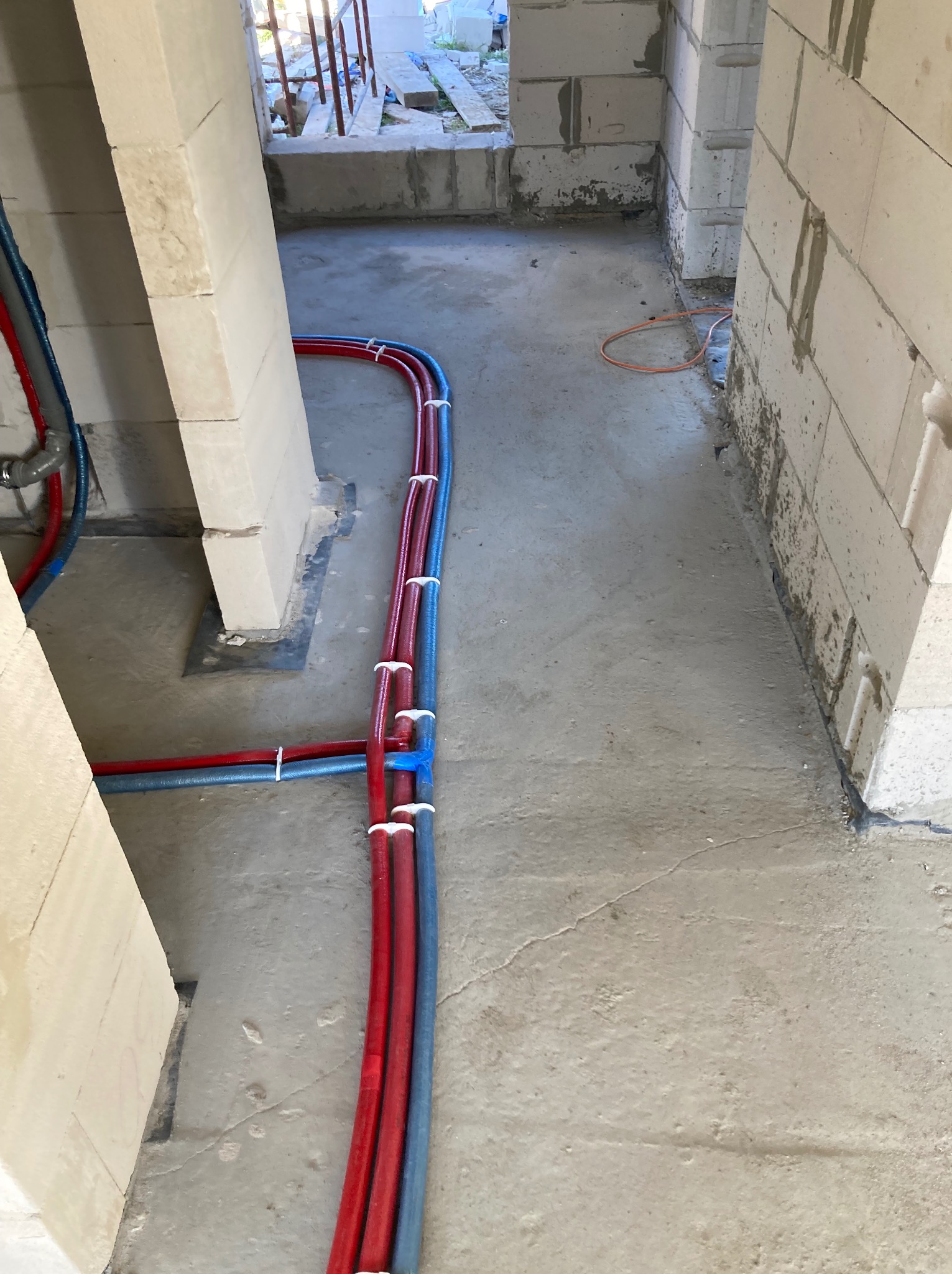 Instalacja rur grzewczych (czerwone i niebieskie) na podłodze w surowym stanie, przymocowane białymi opaskami, widoczne ściany z bloczków betonowych.