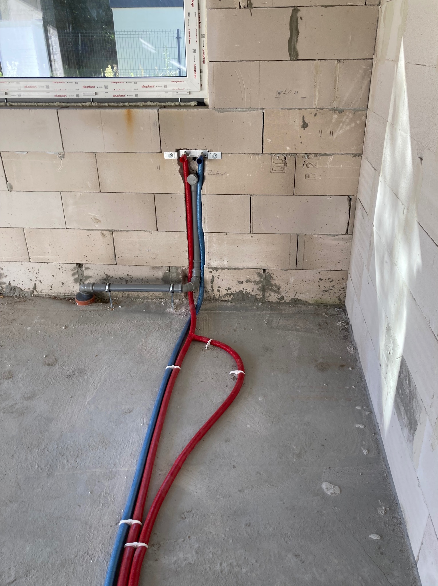 Instalacja hydrauliczna w surowym wnętrzu: czerwone i niebieskie rury prowadzone wzdłuż ściany z betonu komórkowego, widoczne mocowania i fragment okna.