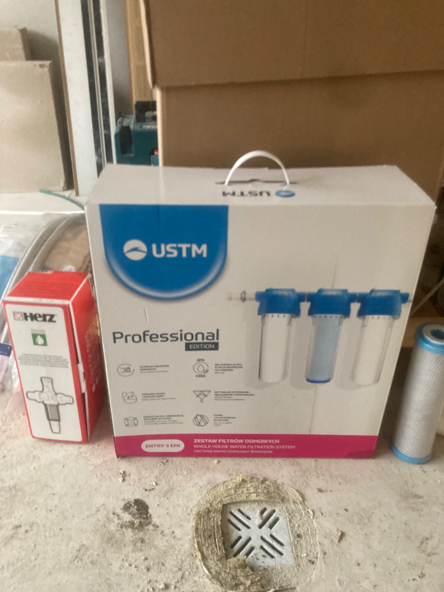 Pudełko z zestawem filtrów domowych USTM Professional Edition, obok zawór Herz i filtr cylindryczny. Widoczna kratka ściekowa w podłodze.