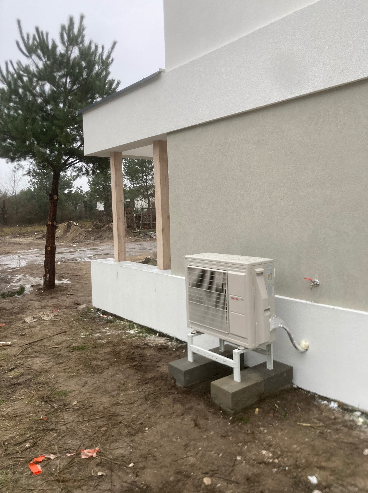 Klimatyzator zewnętrzny zamontowany na betonowych podstawach przy ścianie budynku z widoczną instalacją. Tło stanowi drzewo i teren budowy.