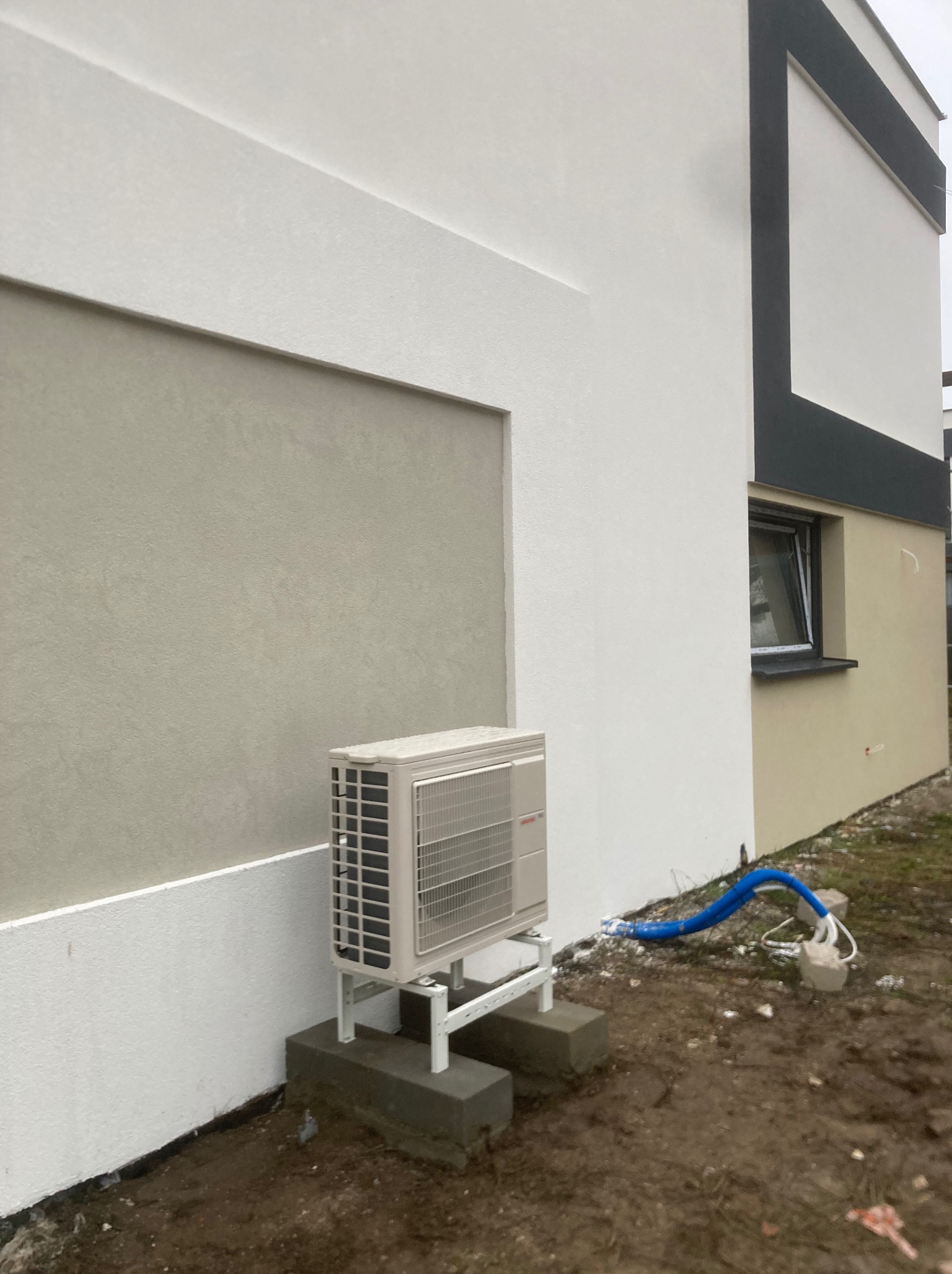 Klimatyzator zewnętrzny zamontowany na betonowych podstawach przy ścianie budynku z niebieskimi rurami odprowadzającymi. Widok z boku, nowoczesna architektura.