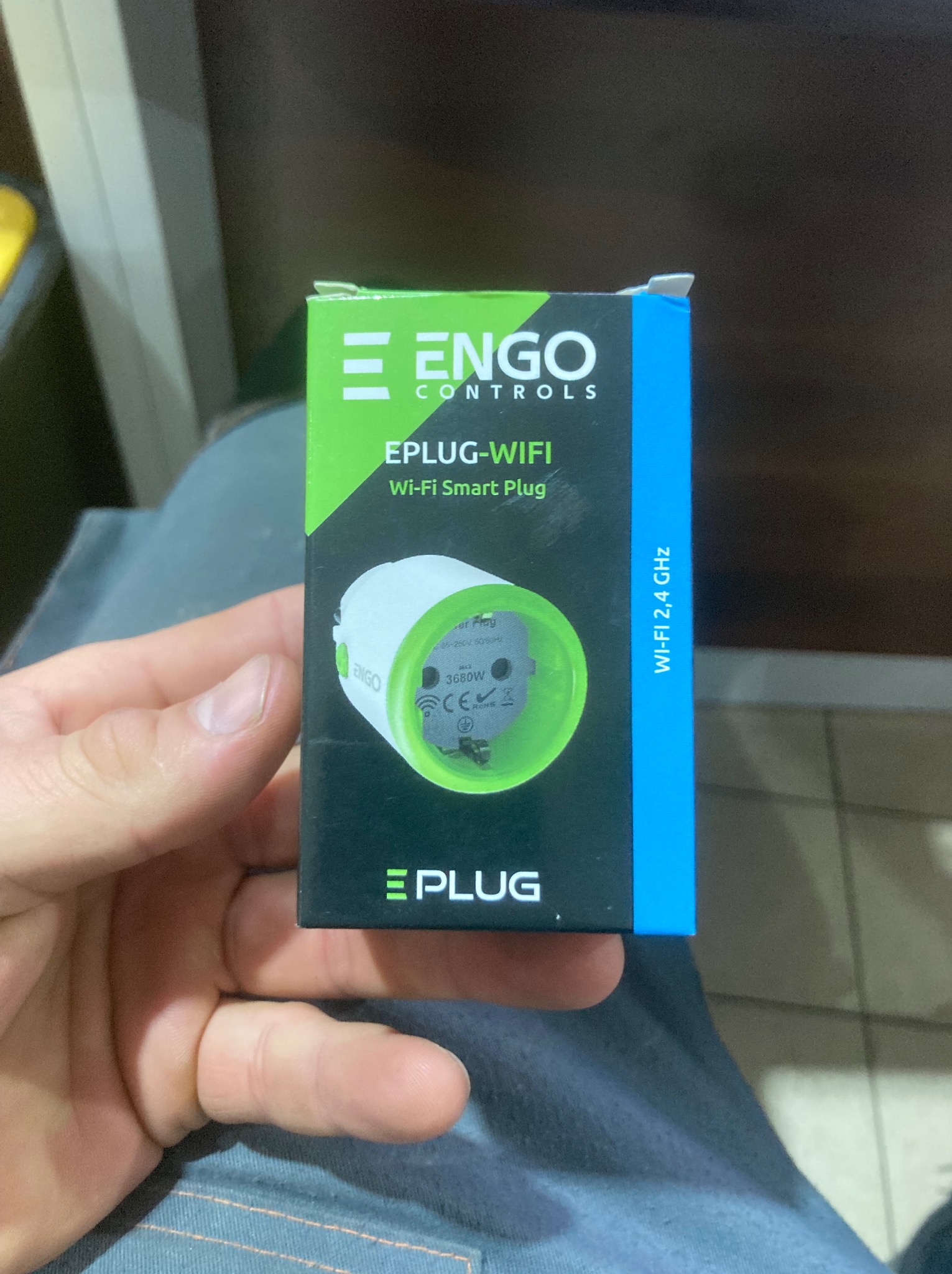 Pudełko z inteligentną wtyczką Wi-Fi ENGO EPlug, trzymane w dłoni, z widocznym nadrukiem specyfikacji i logo. Kolorystyka opakowania: zieleń, czerń i błękit.