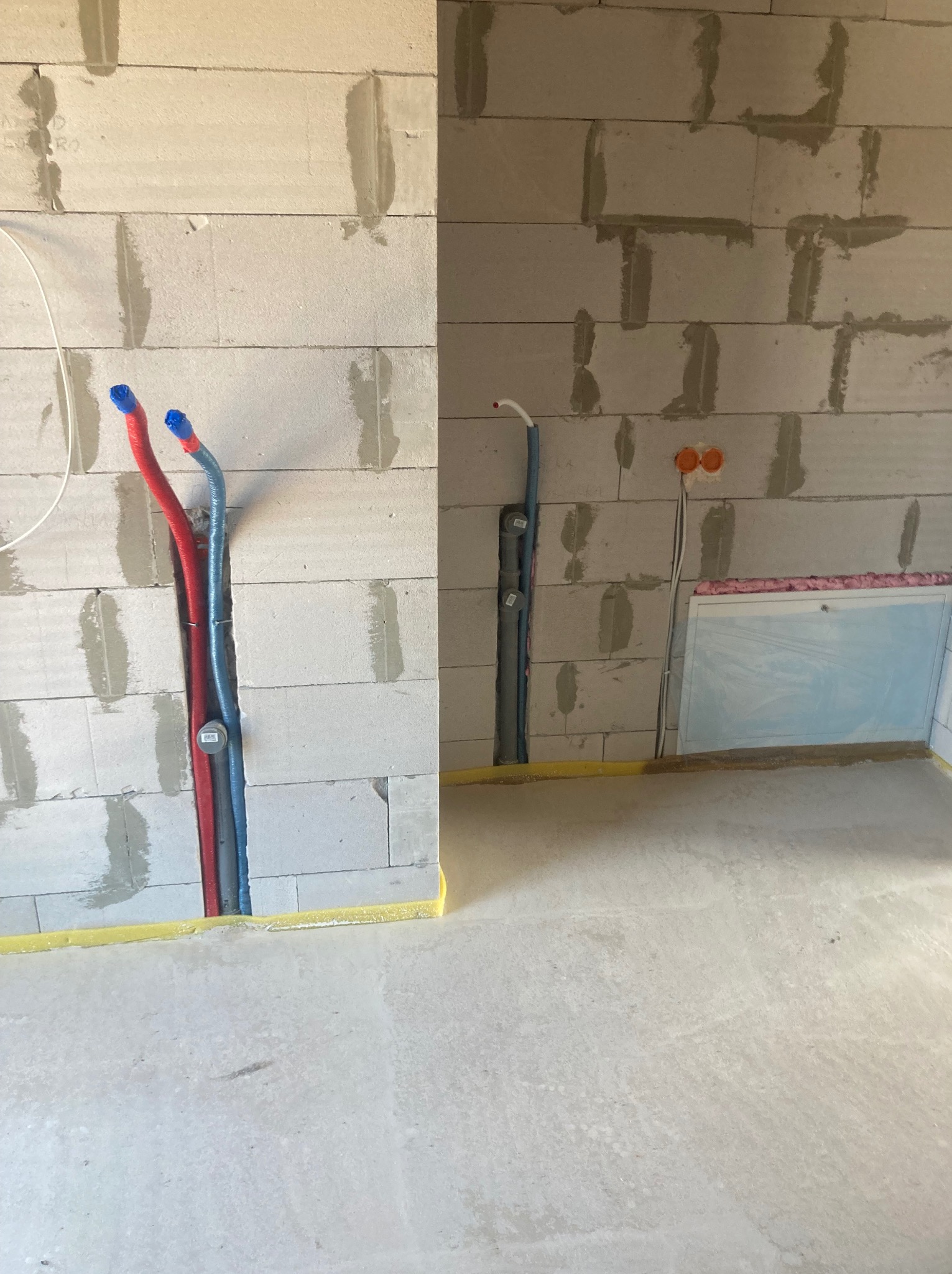 Instalacja hydrauliczna w bruzdach ściany z betonu komórkowego, rury czerwone i niebieskie, żółta taśma zabezpieczająca podłogę, widoczne gniazdka elektryczne.