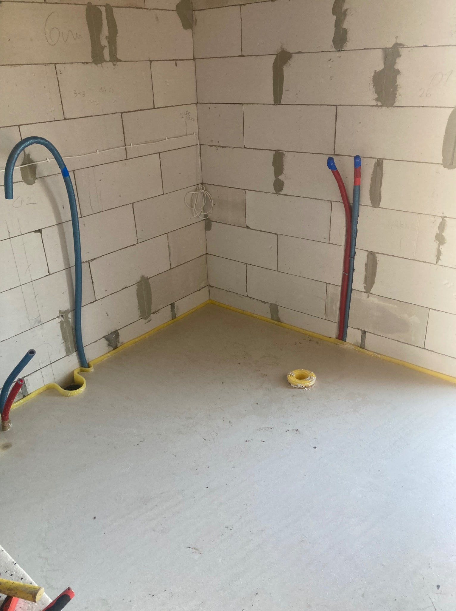 Surowy stan: Instalacja wodna i ogrzewania podłogowego w nowym budynku z bloczków, zabezpieczona żółtą taśmą. Widoczne rury w ścianach i na podłodze, przygotowane do dalszych prac.