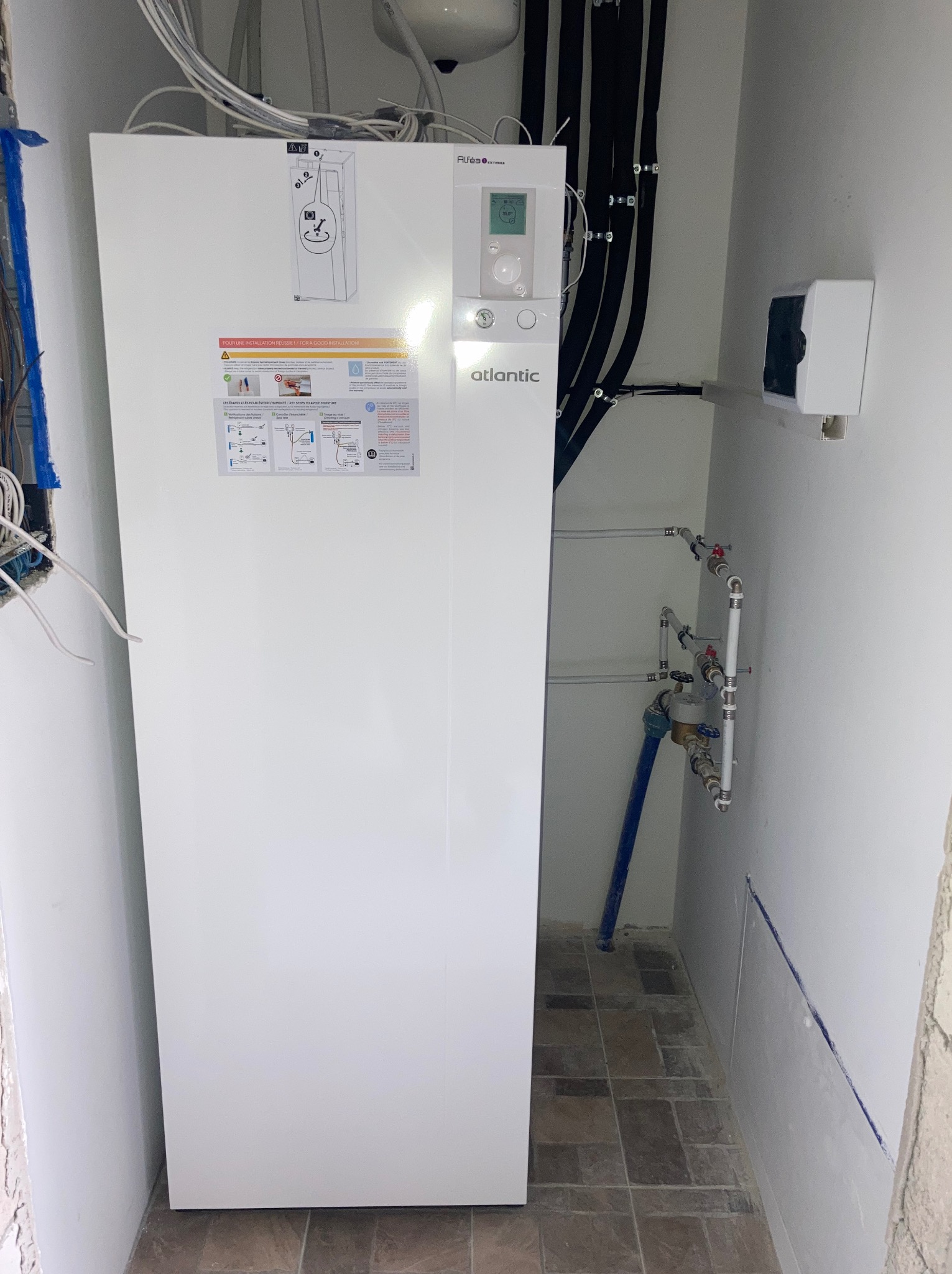 Biała pompa ciepła Atlantic z podłączonymi rurami w pomieszczeniu technicznym, widoczne elementy instalacji hydraulicznej i elektrycznej.