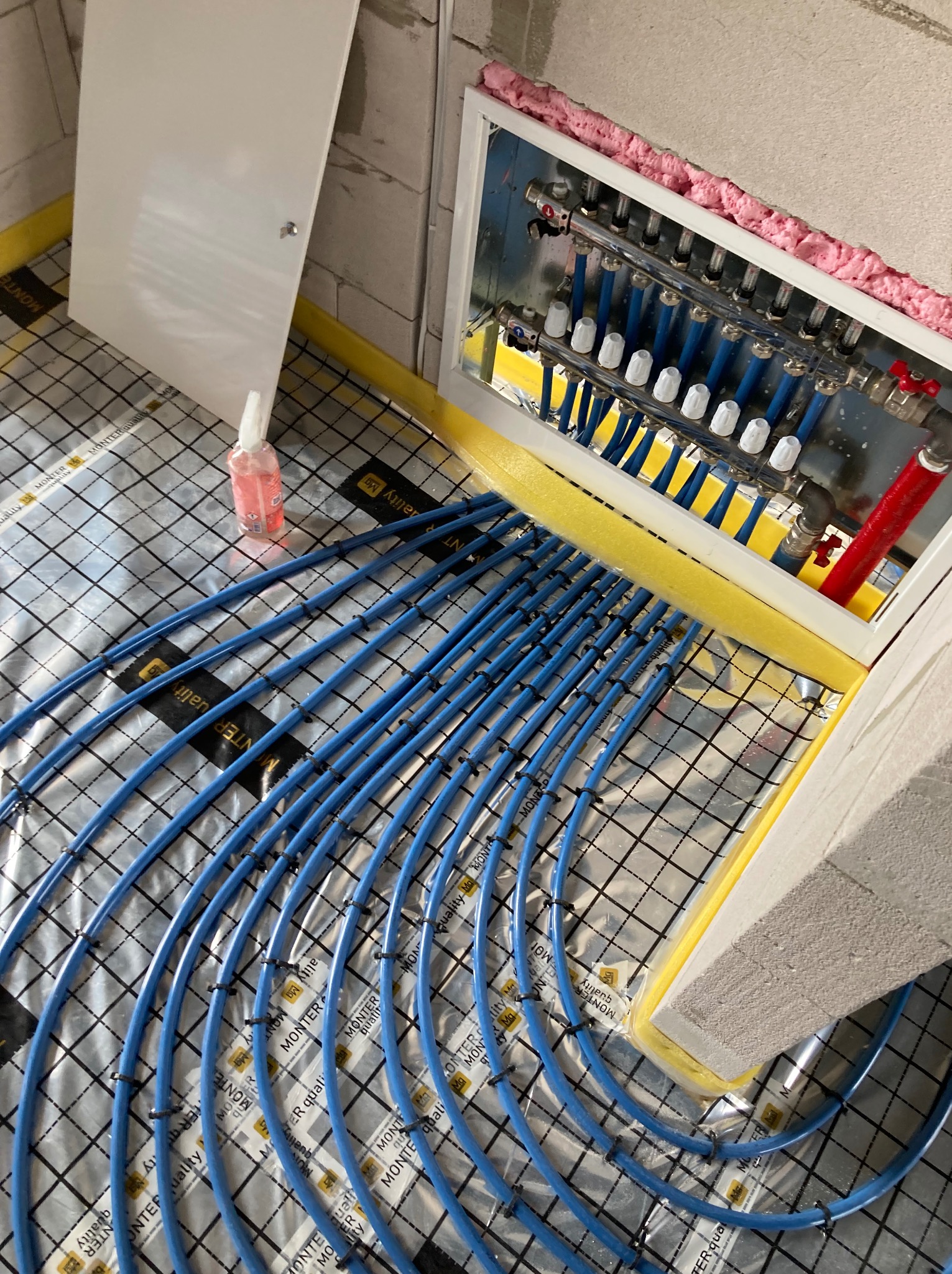 Niebieskie rury ogrzewania podłogowego, przymocowane do folii z nadrukiem, prowadzą do rozdzielacza w białej szafce. Widoczna pianka montażowa i butelka z płynem.