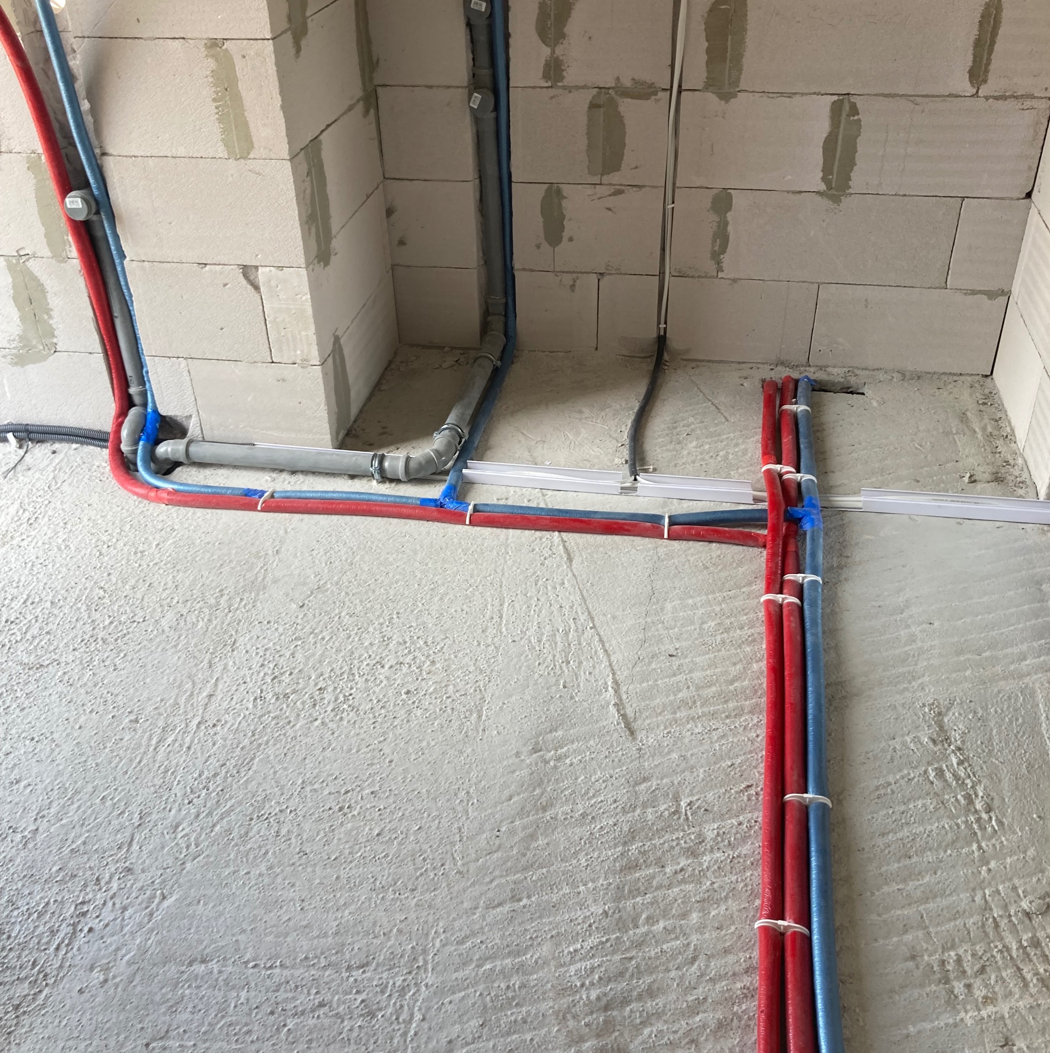 Instalacja wodna i kanalizacyjna w surowym stanie, rury czerwone i niebieskie prowadzone wzdłuż ściany z betonu komórkowego, szare rury kanalizacyjne.