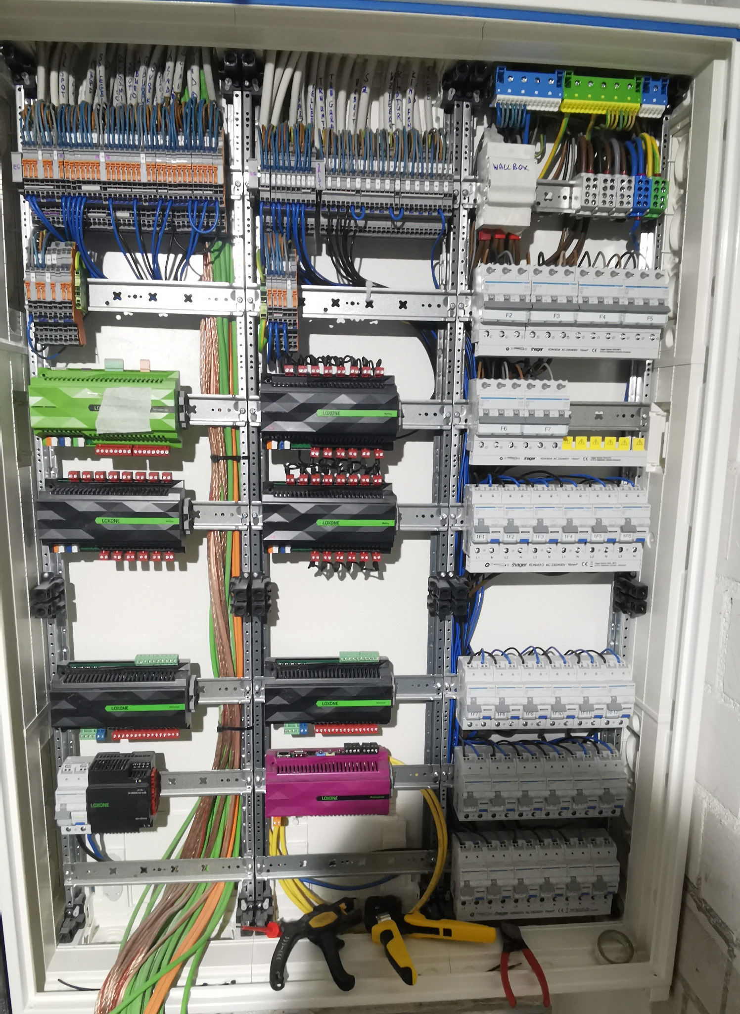 Rozdzielnia elektryczna Loxone z okablowaniem, bezpiecznikami i modułami sterującymi. Widoczne narzędzia: zaciskarka i szczypce. Instalacja w białej obudowie na tle ściany.