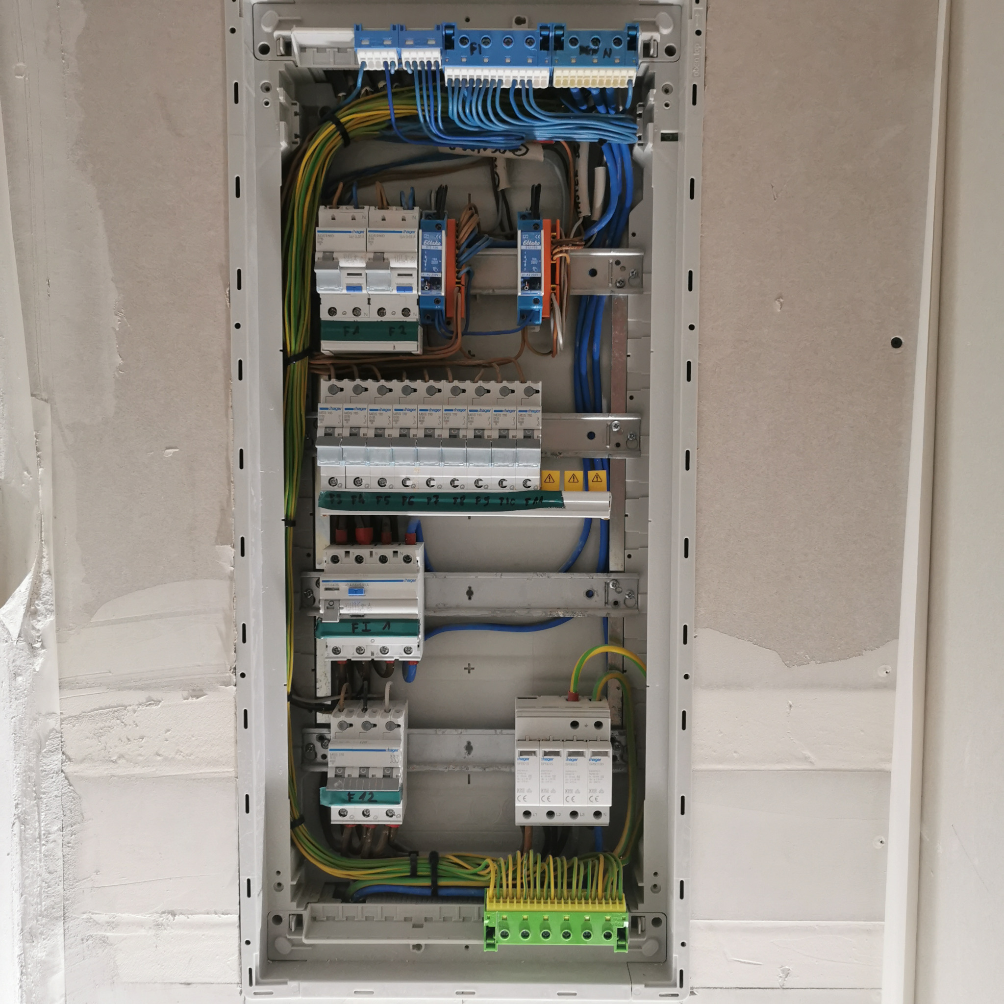 Rozdzielnica elektryczna Hager w szarej obudowie, widoczne zabezpieczenia, kable w kolorach niebieskim, żółto-zielonym, brązowym, zamontowana w ścianie.
