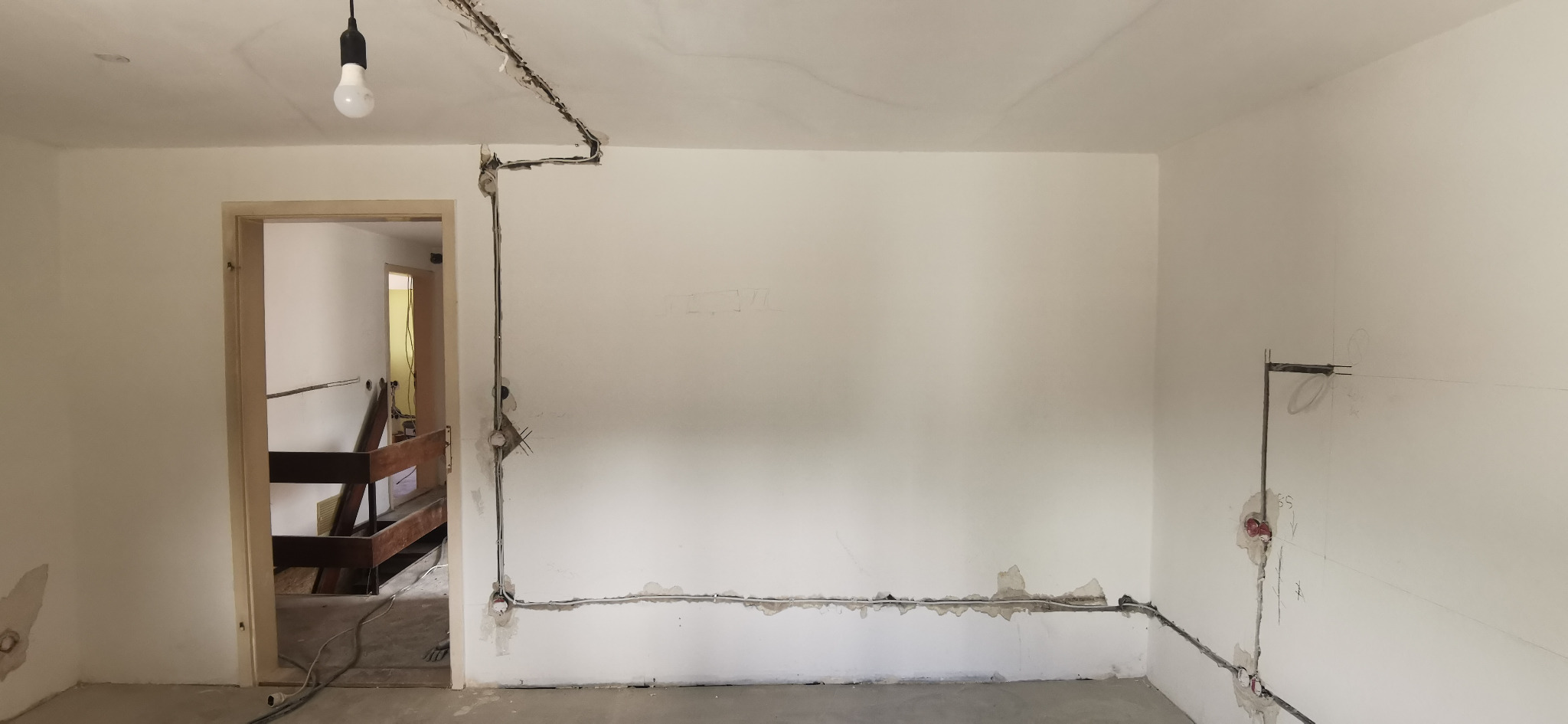 Wnętrze w trakcie prac elektrycznych: kable prowadzone po ścianach, przygotowane punkty instalacyjne. Widoczna surowa ściana i prowizoryczne oświetlenie.