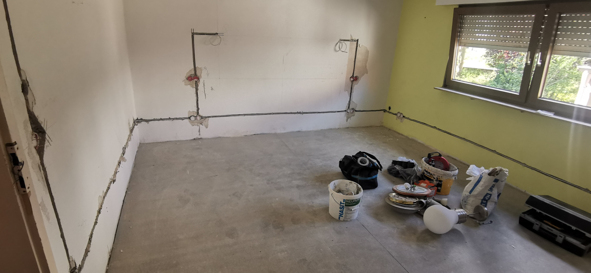 Pokój w trakcie remontu: widoczne przewody elektryczne prowadzone po ścianach, puszki instalacyjne, narzędzia i materiały budowlane na surowej podłodze.