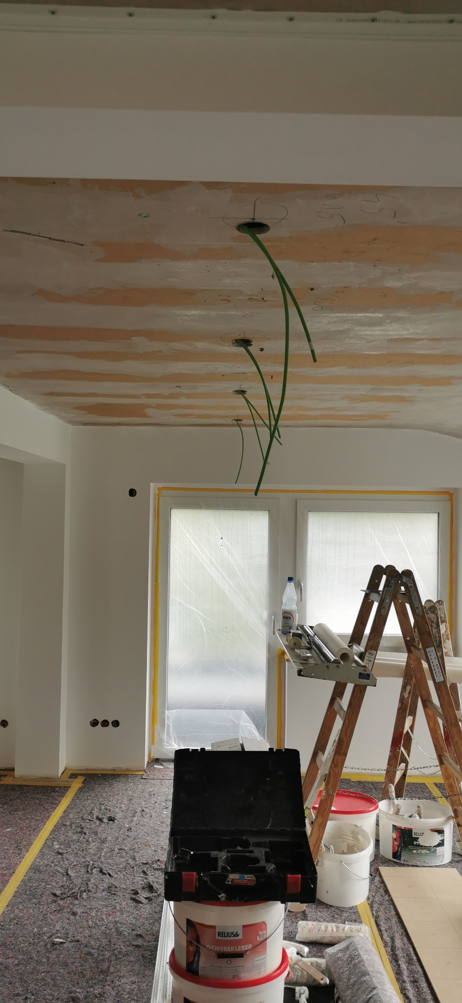 Wnętrze w trakcie remontu: widoczne wystające przewody z sufitu, zabezpieczone okna folią malarską, drabina i narzędzia malarskie.