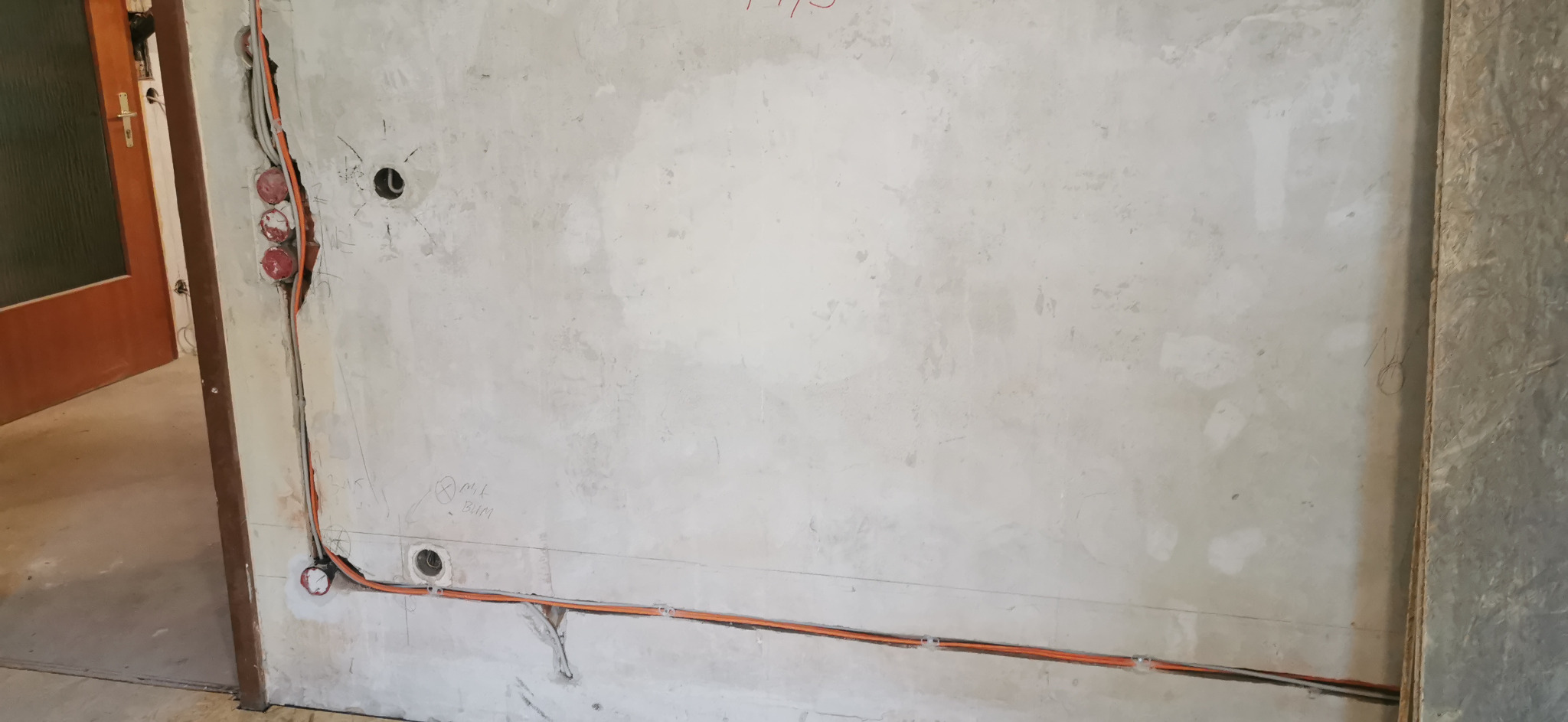 Ściana w trakcie prac instalacyjnych: widoczne przewody elektryczne w korytkach, puszki pod gniazdka, surowy tynk. Etap prac przed wykończeniem.