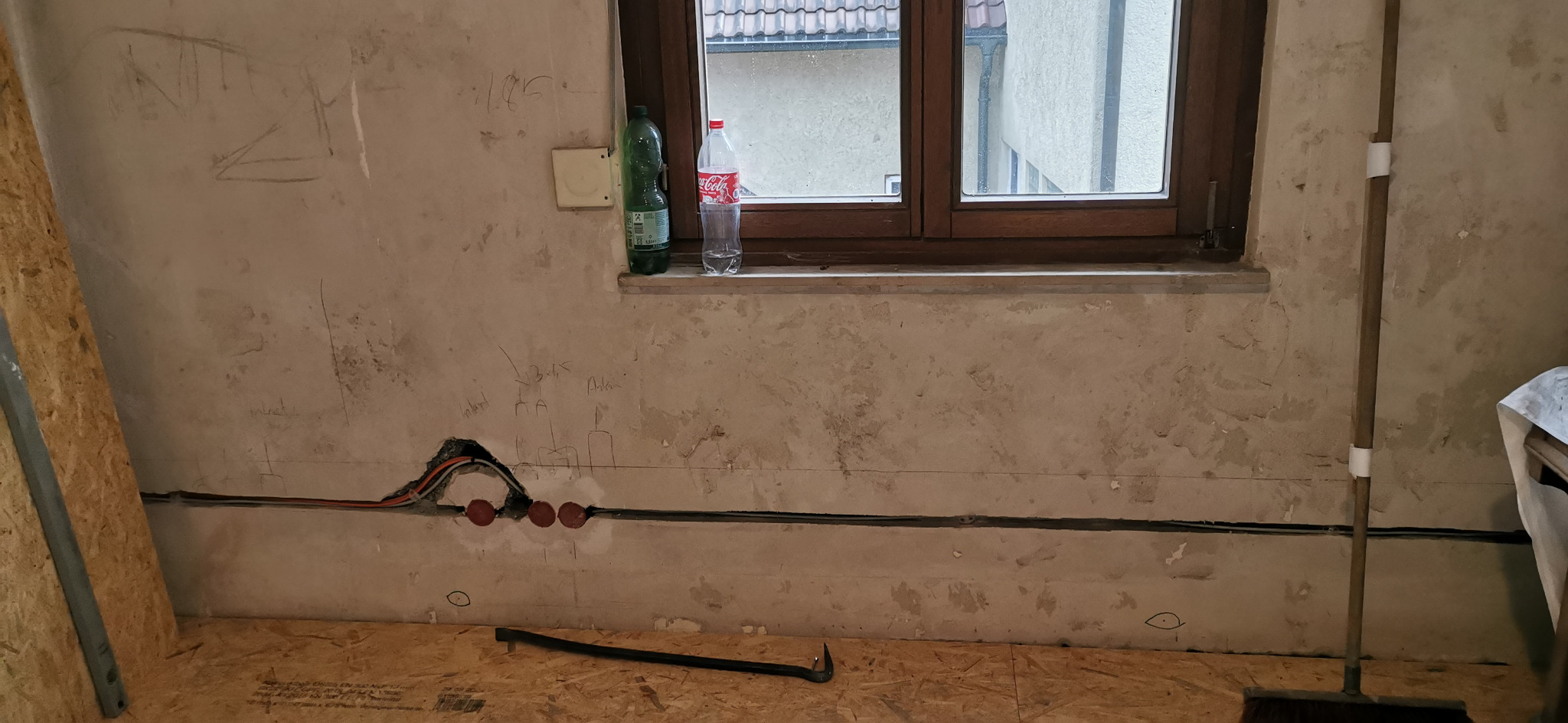 Wnętrze w trakcie remontu: widoczne przewody elektryczne w ścianie z zaznaczonymi punktami instalacji. Na parapecie butelki, na podłodze narzędzie i prowizoryczna podłoga.