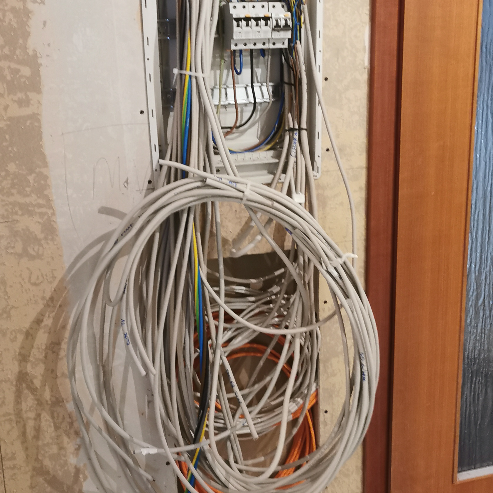 Rozdzielnia elektryczna w trakcie montażu, z widoczną wiązką kabli. Ściana w stanie surowym, obok fragment drewnianej futryny drzwi. Przewody częściowo spięte opaskami.