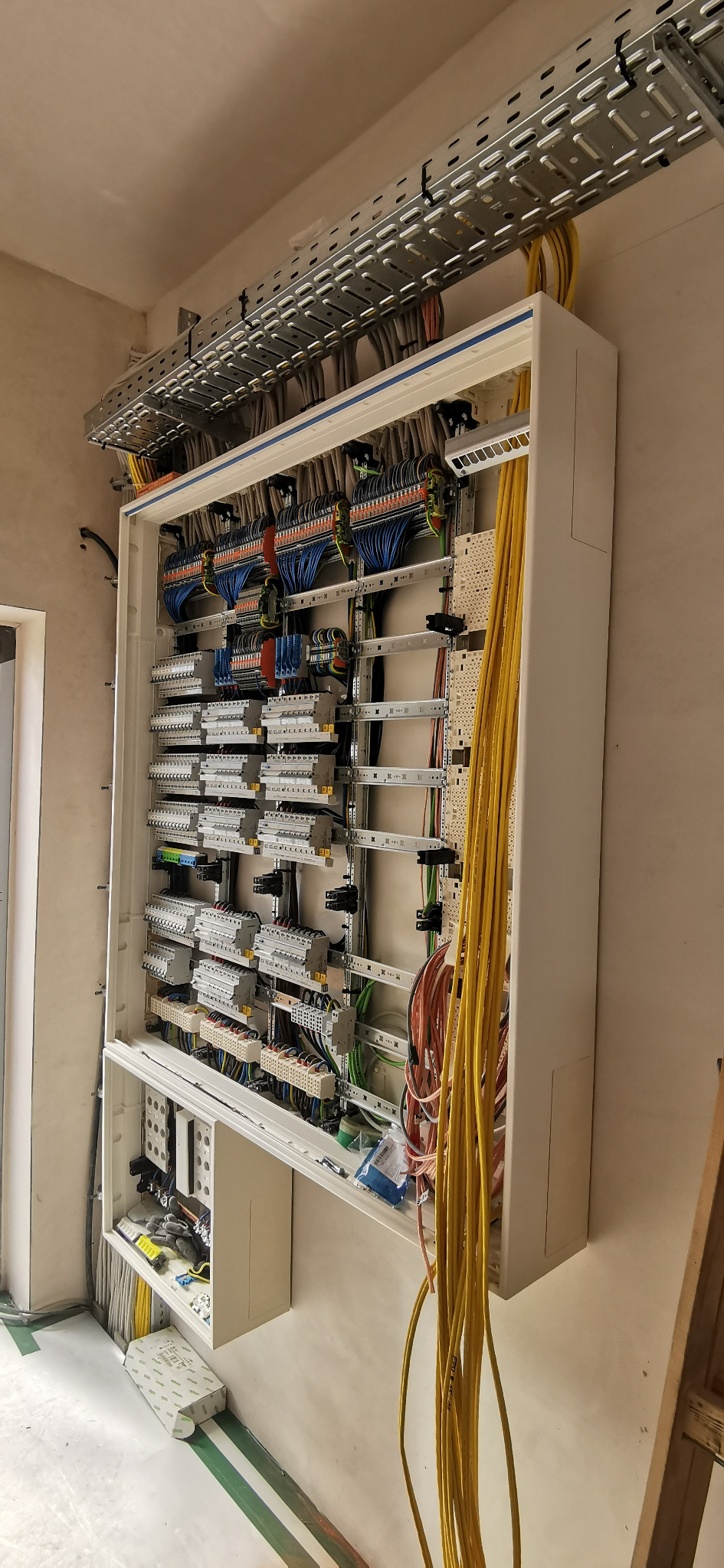 Rozdzielnia elektryczna w białej obudowie, widoczne liczne przewody i zabezpieczenia. Nad rozdzielnią koryto kablowe z ułożonymi przewodami. Ściana w surowym stanie.