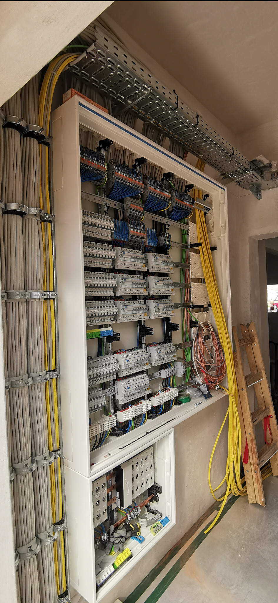 Rozdzielnia elektryczna w trakcie montażu, widoczne kable, zabezpieczenia, drabina i koryto kablowe. Ściany w stanie surowym.