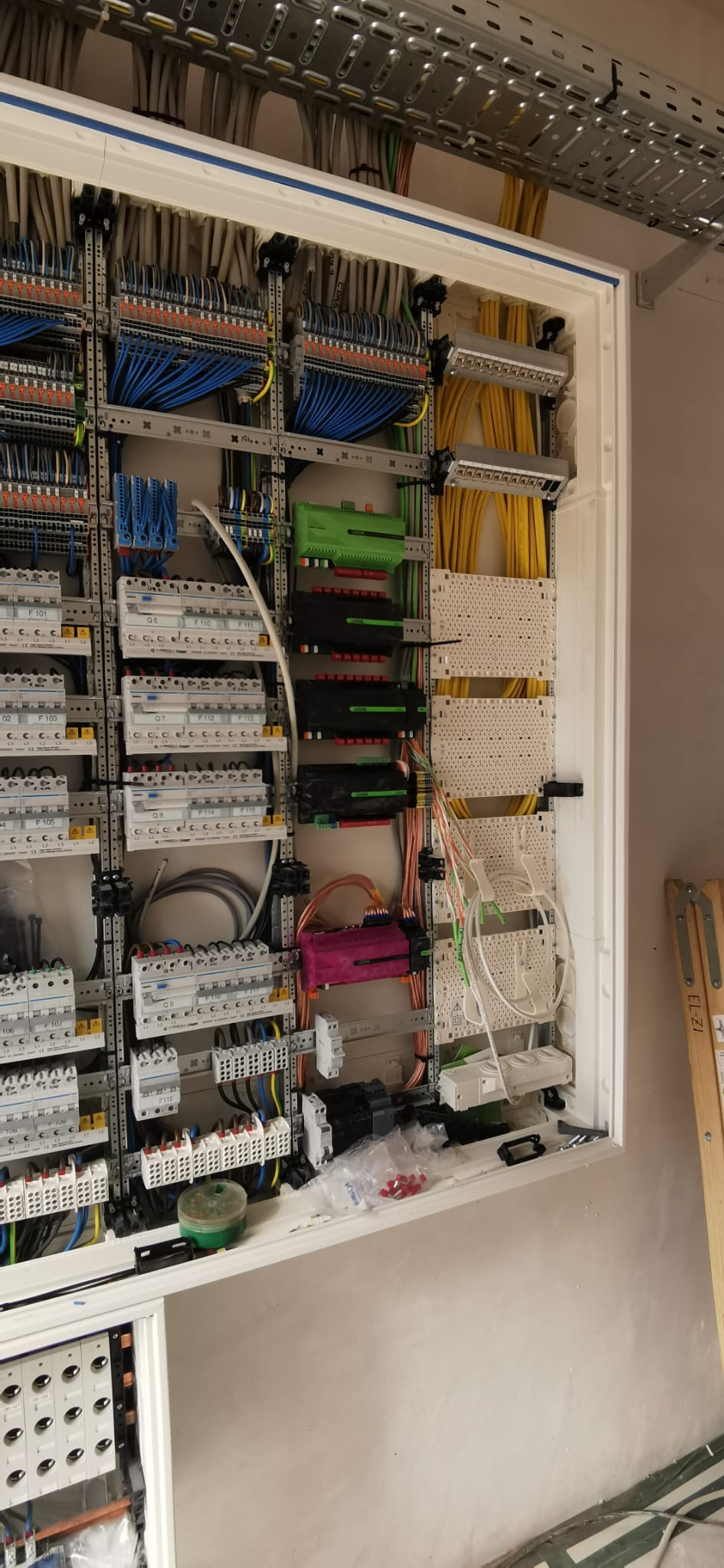 Rozdzielnica elektryczna z precyzyjnie ułożonymi kablami w różnych kolorach (niebieskie, żółte) i modułami. Częściowo widoczne akcesoria i narzędzia.