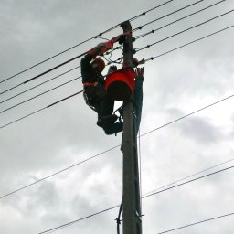 PRO-ELECTRO MATEUSZ BORAWSKI - Dwóch monterów w kaskach i uprzężach pracuje na słupie energetycznym pod pochmurnym niebem. Widoczne linie wysokiego napięcia i sprzęt roboczy.