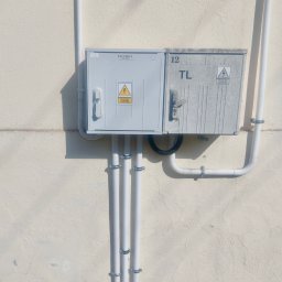 PRO-ELECTRO MATEUSZ BORAWSKI - Szara skrzynka z oznaczeniami ostrzegawczymi przymocowana do ściany. Widoczne białe rury zabezpieczone metalowymi obejmami. Element instalacji elektrycznej na zewnątrz budynku.