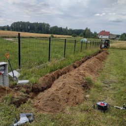 PRO-ELECTRO MATEUSZ BORAWSKI - Wykopany rów wzdłuż ogrodzenia, widoczna skrzynka elektryczna, narzędzia, mini koparka w tle. Prace ziemne przygotowujące do instalacji podziemnej.