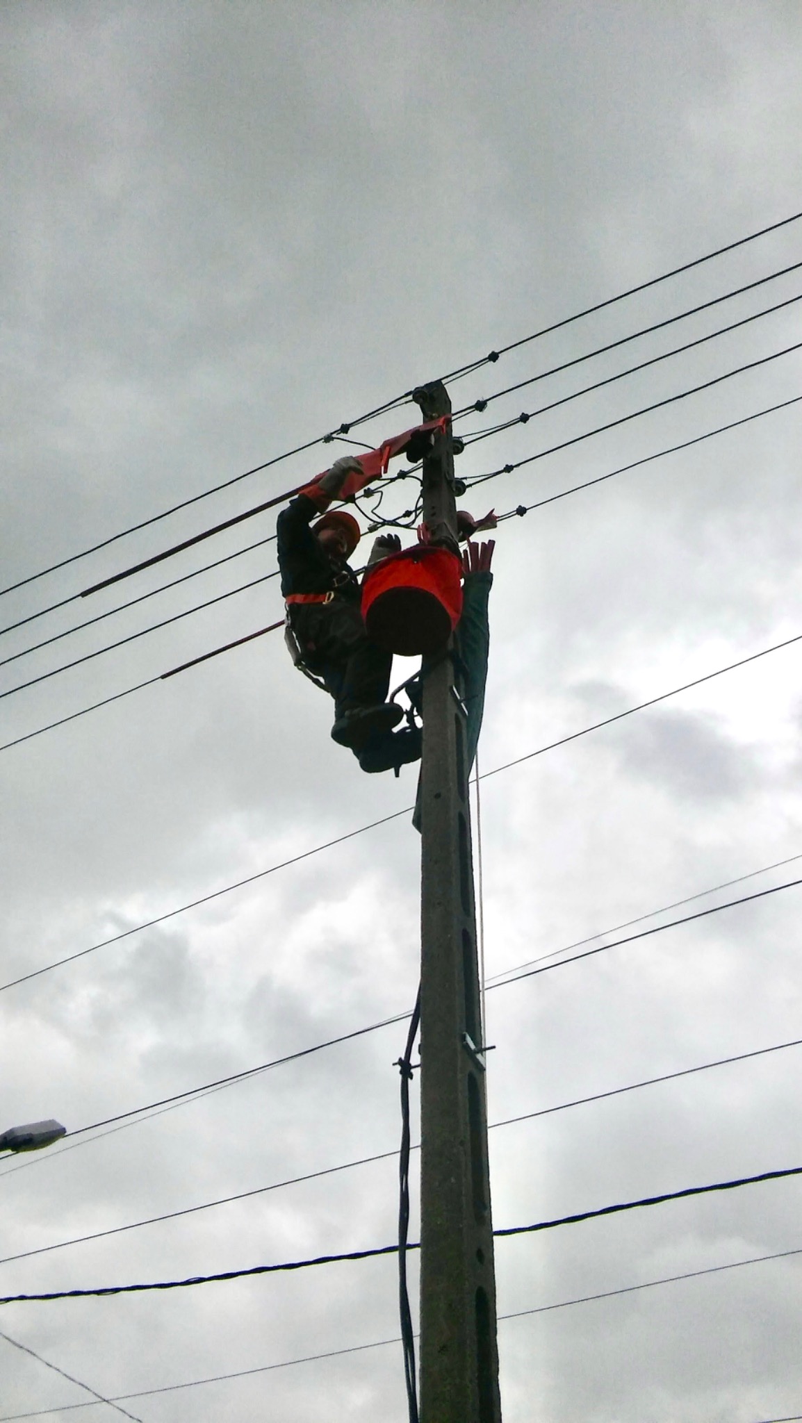 Dwóch monterów w kaskach i uprzężach pracuje na słupie energetycznym pod pochmurnym niebem. Widoczne linie wysokiego napięcia i sprzęt roboczy.