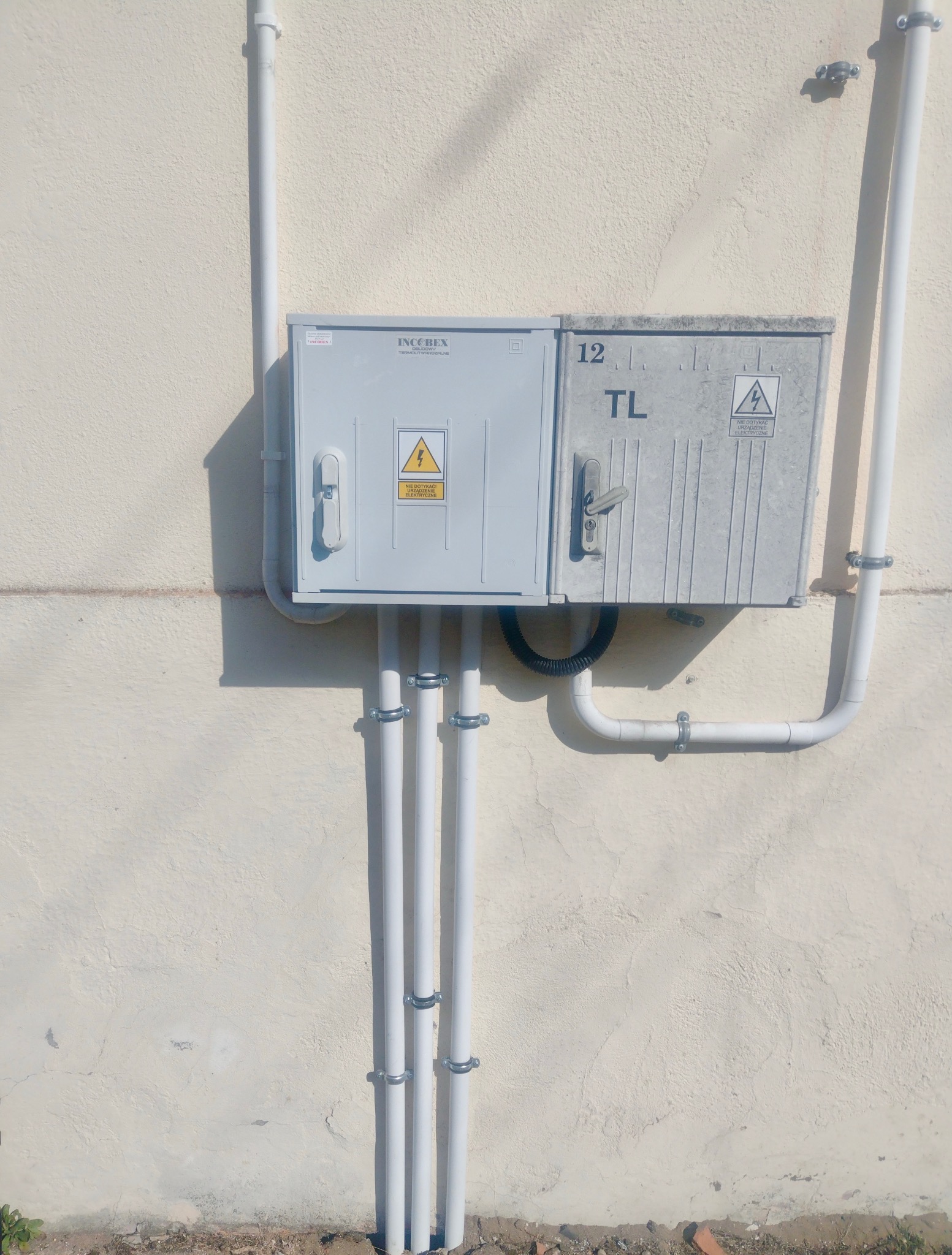 Szara skrzynka z oznaczeniami ostrzegawczymi przymocowana do ściany. Widoczne białe rury zabezpieczone metalowymi obejmami. Element instalacji elektrycznej na zewnątrz budynku.