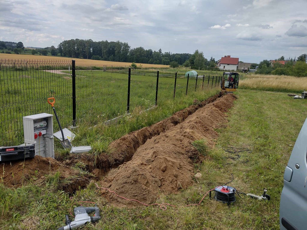 Wykopany rów wzdłuż ogrodzenia, widoczna skrzynka elektryczna, narzędzia, mini koparka w tle. Prace ziemne przygotowujące do instalacji podziemnej.