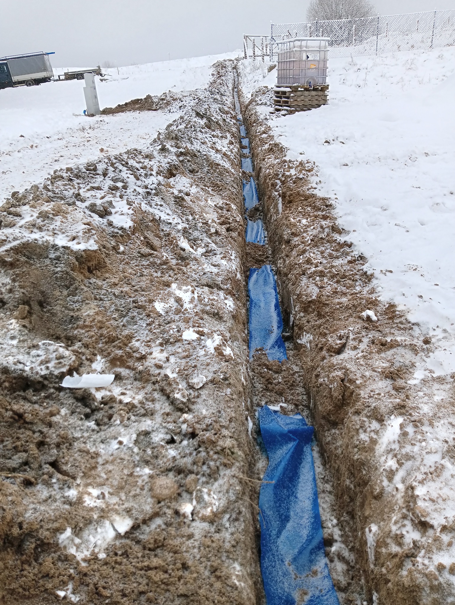 Wykop w ziemi, wyłożony niebieską folią, prawdopodobnie pod instalację. Śnieżny krajobraz w tle. Widoczny zbiornik na paletach i fragment ogrodzenia.