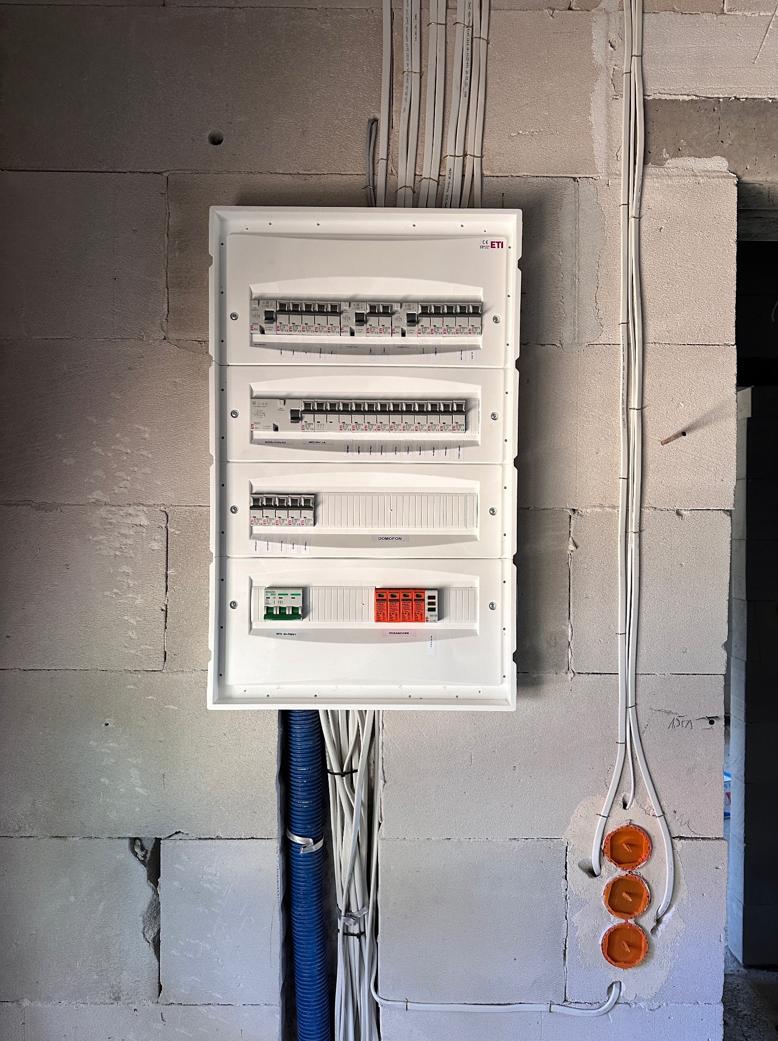 Rozdzielnica elektryczna zamontowana na surowej ścianie z betonu komórkowego, widoczne kable i peszel. Instalacja w trakcie realizacji.