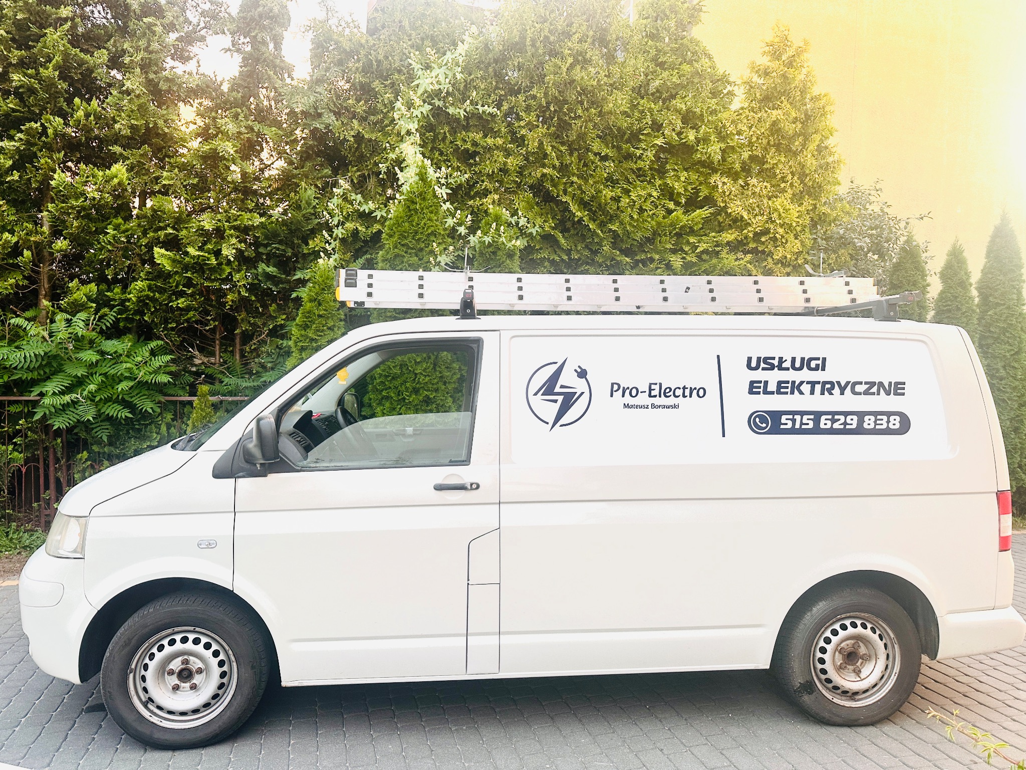 Biały samochód dostawczy z logo i numerem telefonu firmy elektrycznej 'Pro-Electro' na tle zielonych drzew i drabiny na dachu.