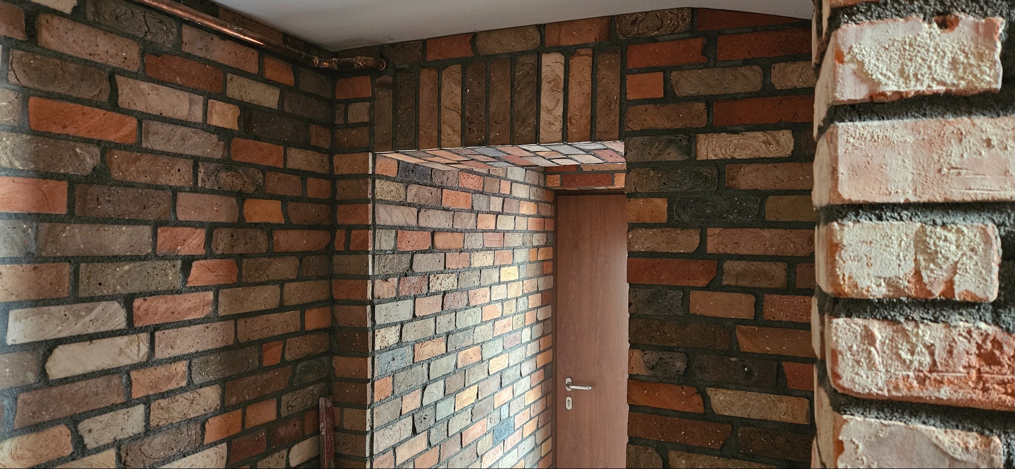 Wnętrze z dekoracyjną ścianą z cegły w odcieniach brązu i czerwieni, z widocznym wejściem do pomieszczenia i fragmentem instalacji miedzianej pod sufitem.