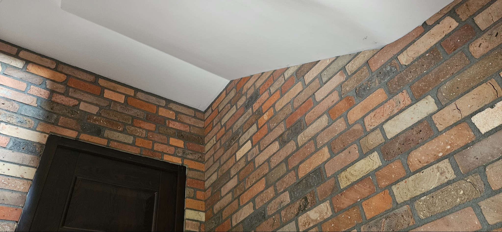 Ściana z dekoracyjnej cegły w odcieniach brązu i beżu, połączona z białym sufitem i fragmentem ciemnych drzwi. Widok na róg pomieszczenia.