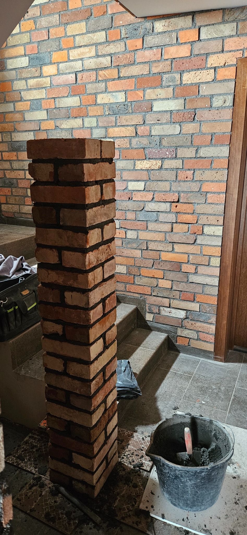 Wznoszenie ceglanego filaru z użyciem zaprawy, widoczny fragment ściany z cegieł w tle, wiadro z zaprawą i narzędzia na podłodze, etap prac murarskich.
