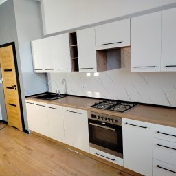 Wykończenia wnętrz i remonty mieszkań - Nowoczesna kuchnia z białymi szafkami, drewnianym blatem i marmurowym panelem ściennym. Widoczny zlew, płyta gazowa i piekarnik. Jasne drewniane panele na podłodze.