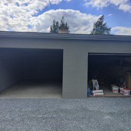 Wykończenia wnętrz i remonty mieszkań - Szary, otynkowany garaż dwustanowiskowy z otwartymi wrotami, widok z zewnątrz na żwirowe podłoże. Na wrotach widoczne worki z zaprawą i puste wiadra po farbie.