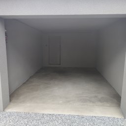 Wykończenia wnętrz i remonty mieszkań - Wnętrze garażu z szarą, teksturowaną ścianą i świeżo wylaną, gładką posadzką. Na zewnątrz widoczne kamienie i rura spustowa.