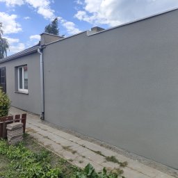 Wykończenia wnętrz i remonty mieszkań - Ściana budynku po tynkowaniu elewacji w szarym kolorze z widoczną rynną, okno z brązowymi okiennicami i fragmentem chodnika z kostki. Budynek jednokondygnacyjny.