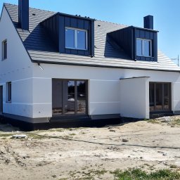 Wykończenia wnętrz i remonty mieszkań - Nowoczesny dom z szarą dachówką i białymi ścianami, widoczny z zewnątrz. Budynek z oknami i kominami, otoczony placem budowy. Częściowo ukończona elewacja.