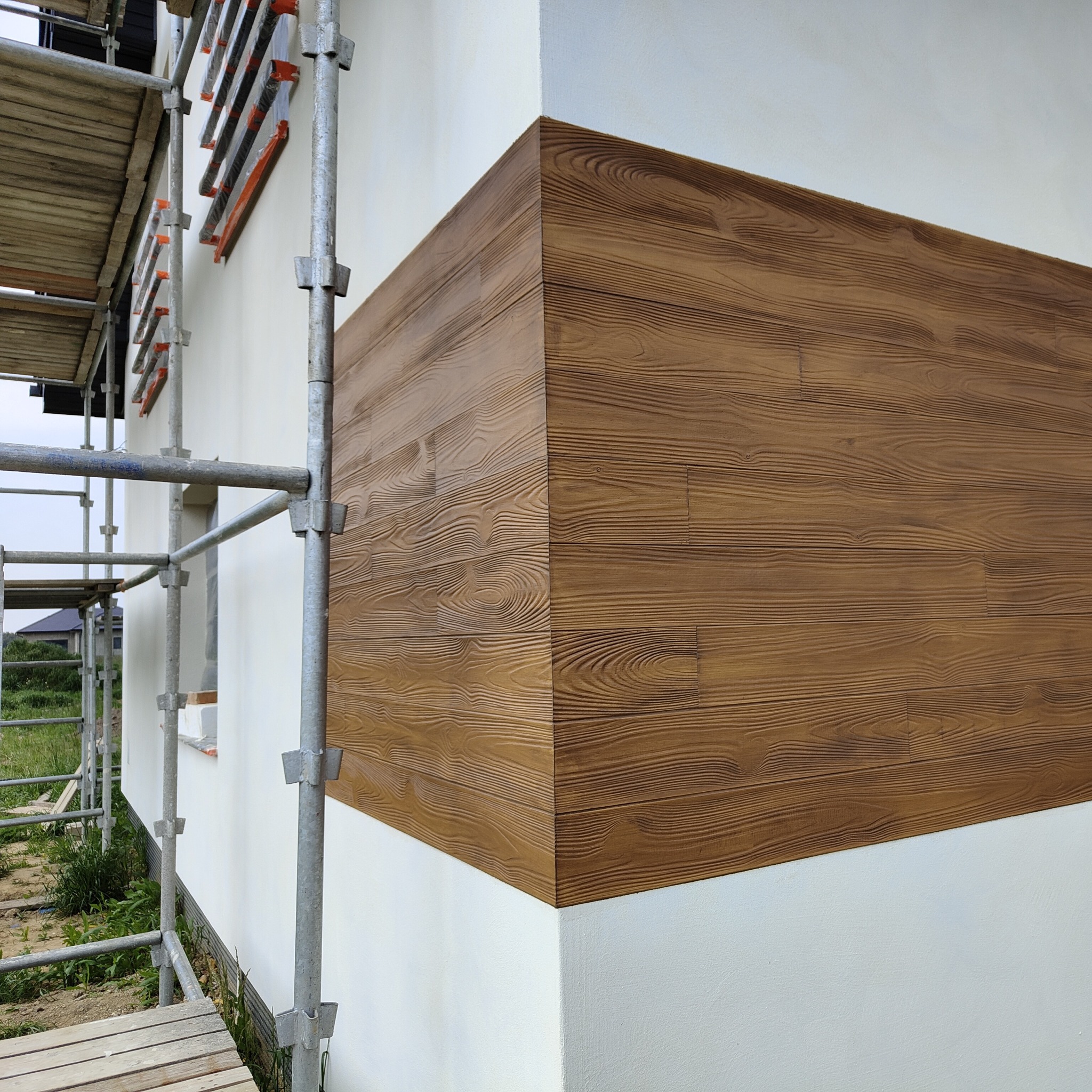 Wykończona elewacja budynku z dekoracyjnym panelem imitującym drewno, widoczne rusztowanie. Detal narożnika i faktury paneli w ciepłym odcieniu brązu.