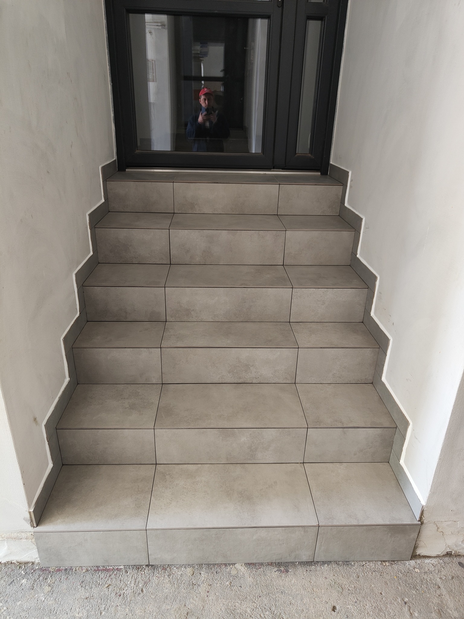 Wykończone płytkami schody wejściowe z szarą fugą i widocznym odbiciem osoby w szybie drzwi wejściowych. Minimalistyczne wykończenie.
