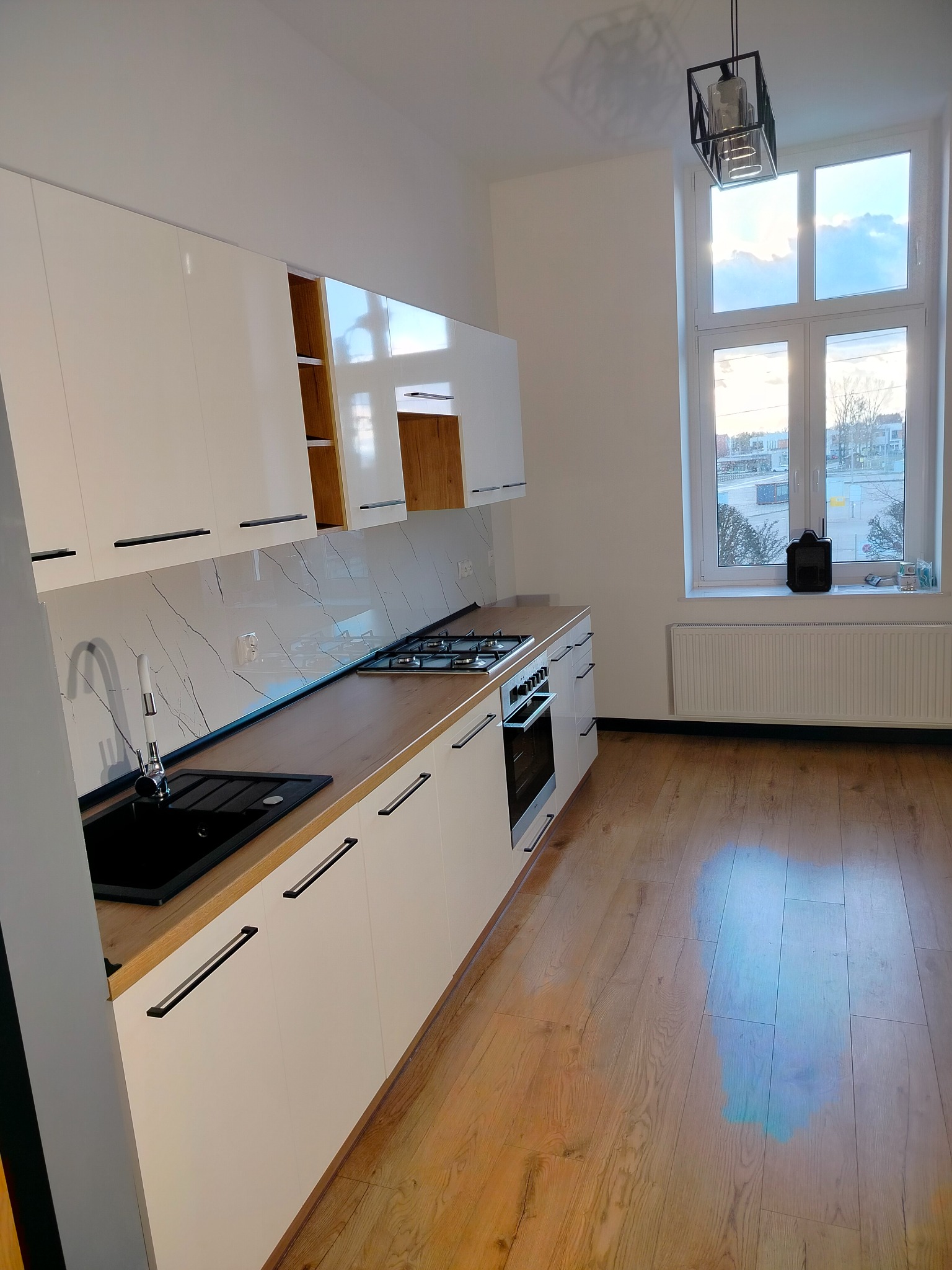 Biała kuchnia z drewnianym blatem i czarnymi uchwytami w Małgowie. Widok na okno z widokiem na zewnątrz. Nowoczesne wykończenie i jasne wnętrze.