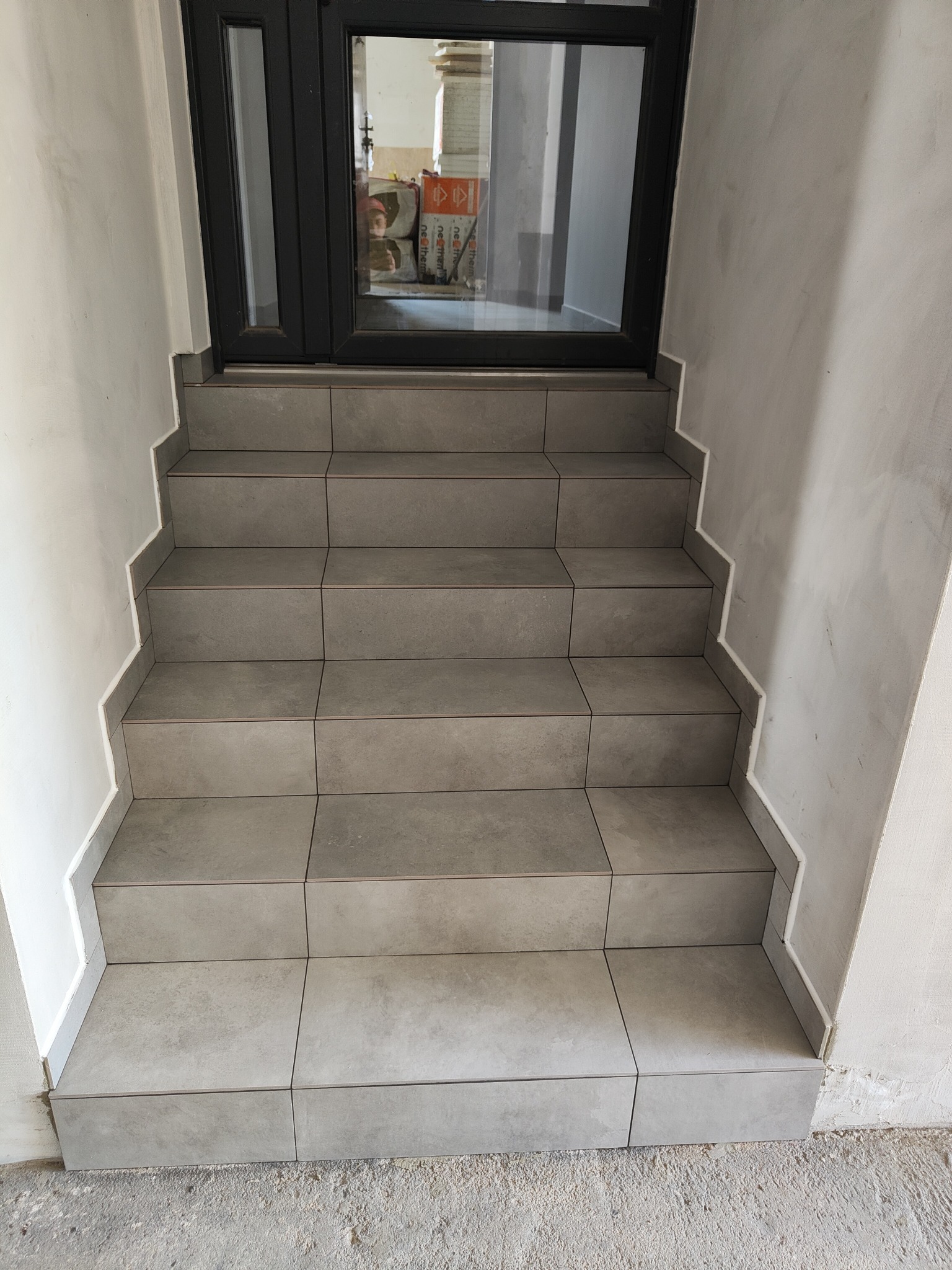 Wykończone schody wejściowe z szarych płytek, z widocznym oknem/drzwiami wejściowymi. Płytki ułożone starannie, z fugami w kolorze białym.