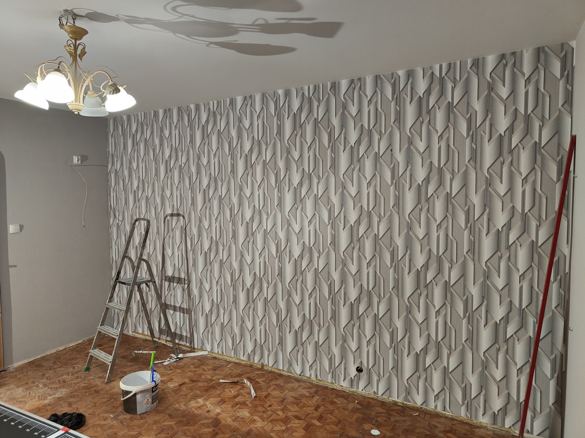 Ściana z wzorzystą tapetą w odcieniach szarości, drabina malarska i akcesoria. Parkiet z klepek. Wnętrze w trakcie prac wykończeniowych.