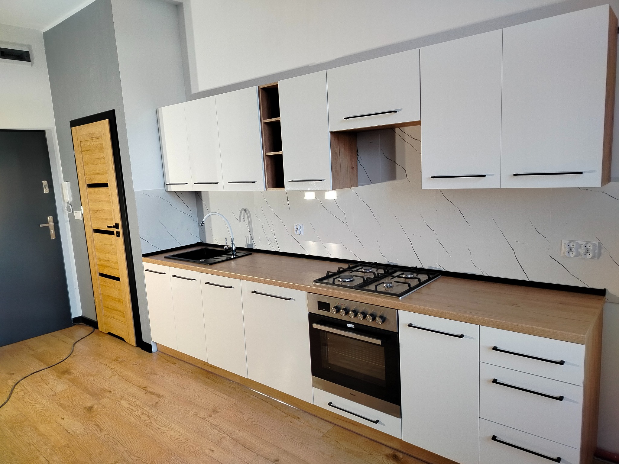 Nowoczesna kuchnia z białymi szafkami, drewnianym blatem i marmurowym panelem ściennym. Widoczny zlew, płyta gazowa i piekarnik. Jasne drewniane panele na podłodze.