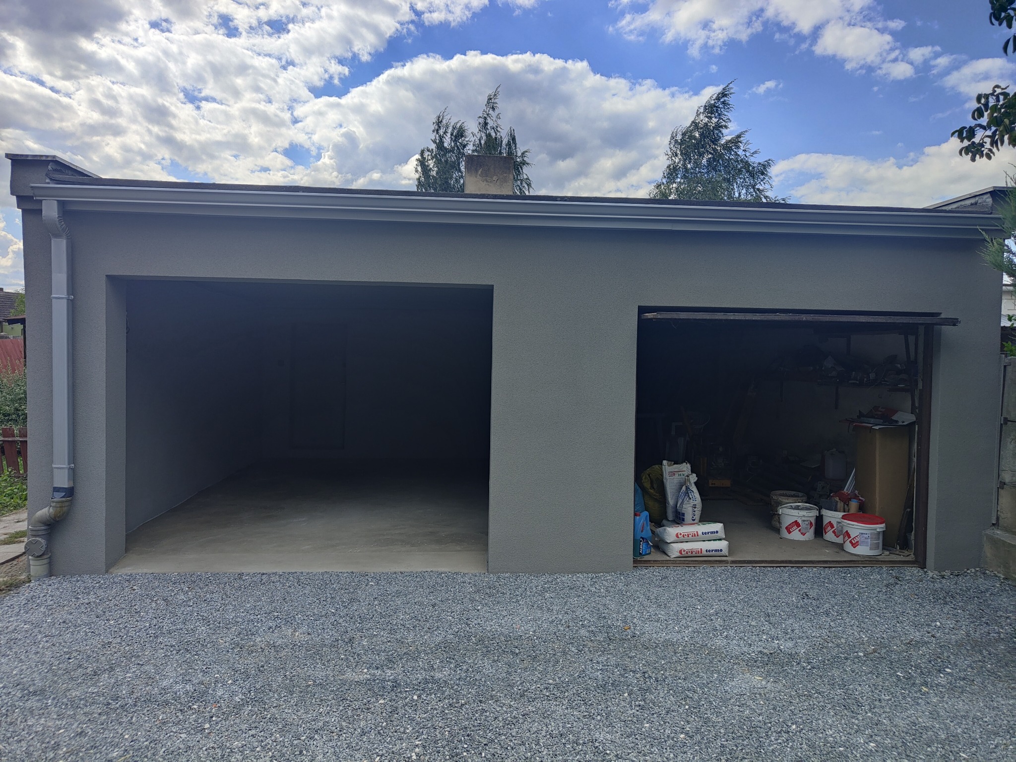 Szary, otynkowany garaż dwustanowiskowy z otwartymi wrotami, widok z zewnątrz na żwirowe podłoże. Na wrotach widoczne worki z zaprawą i puste wiadra po farbie.