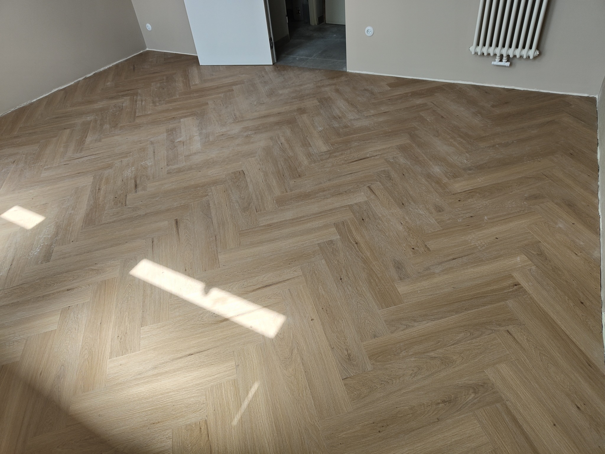 Ukończona podłoga z paneli ułożonych w jodełkę, oświetlona naturalnym światłem słonecznym, w tle biały grzejnik i drzwi.