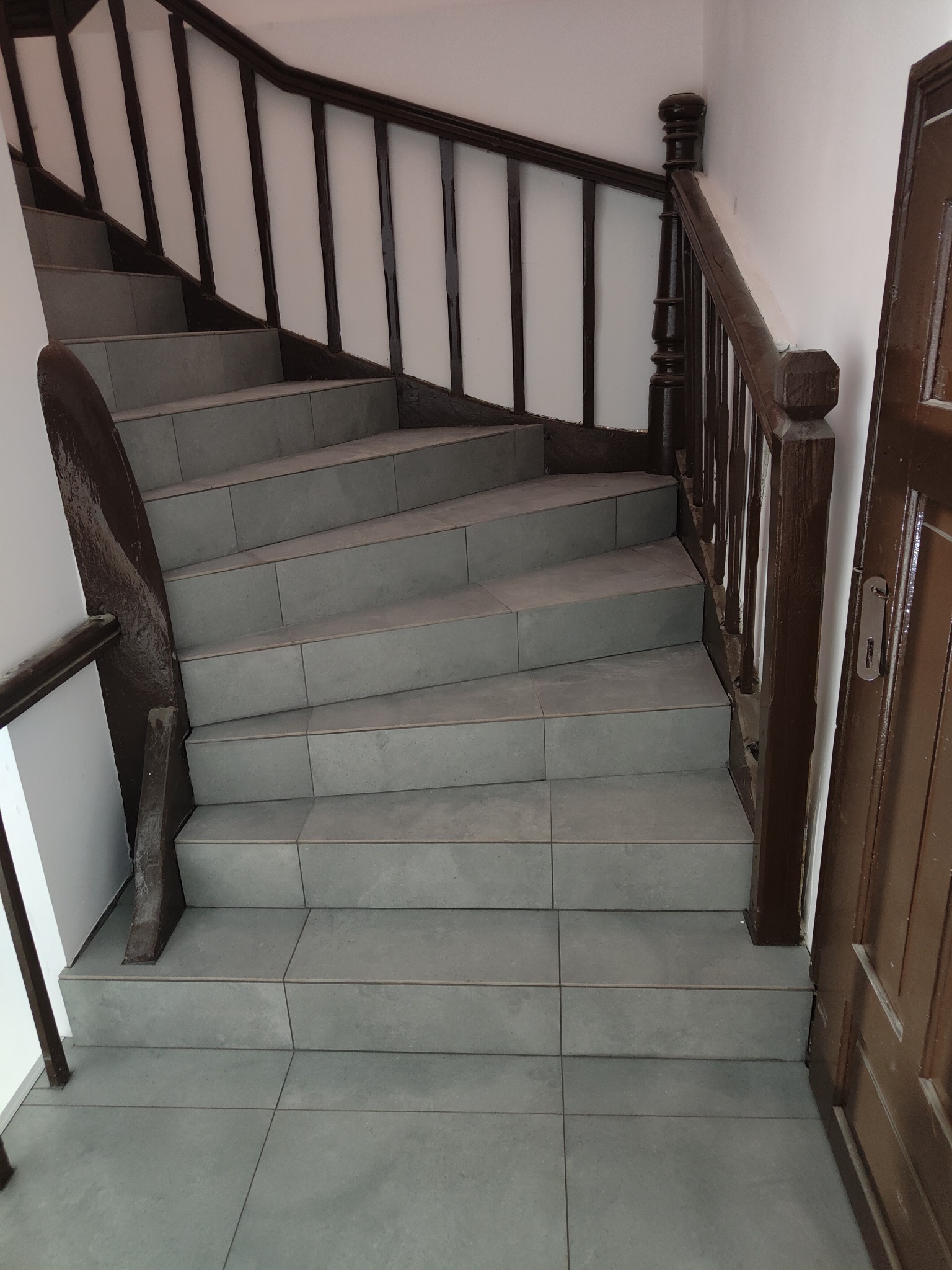 Wykończone schody z szarych płytek, ciemnobrązowa balustrada i poręcz. Widoczne fragmenty brązowych drzwi. Ujęcie z góry, podkreślające symetrię stopni.