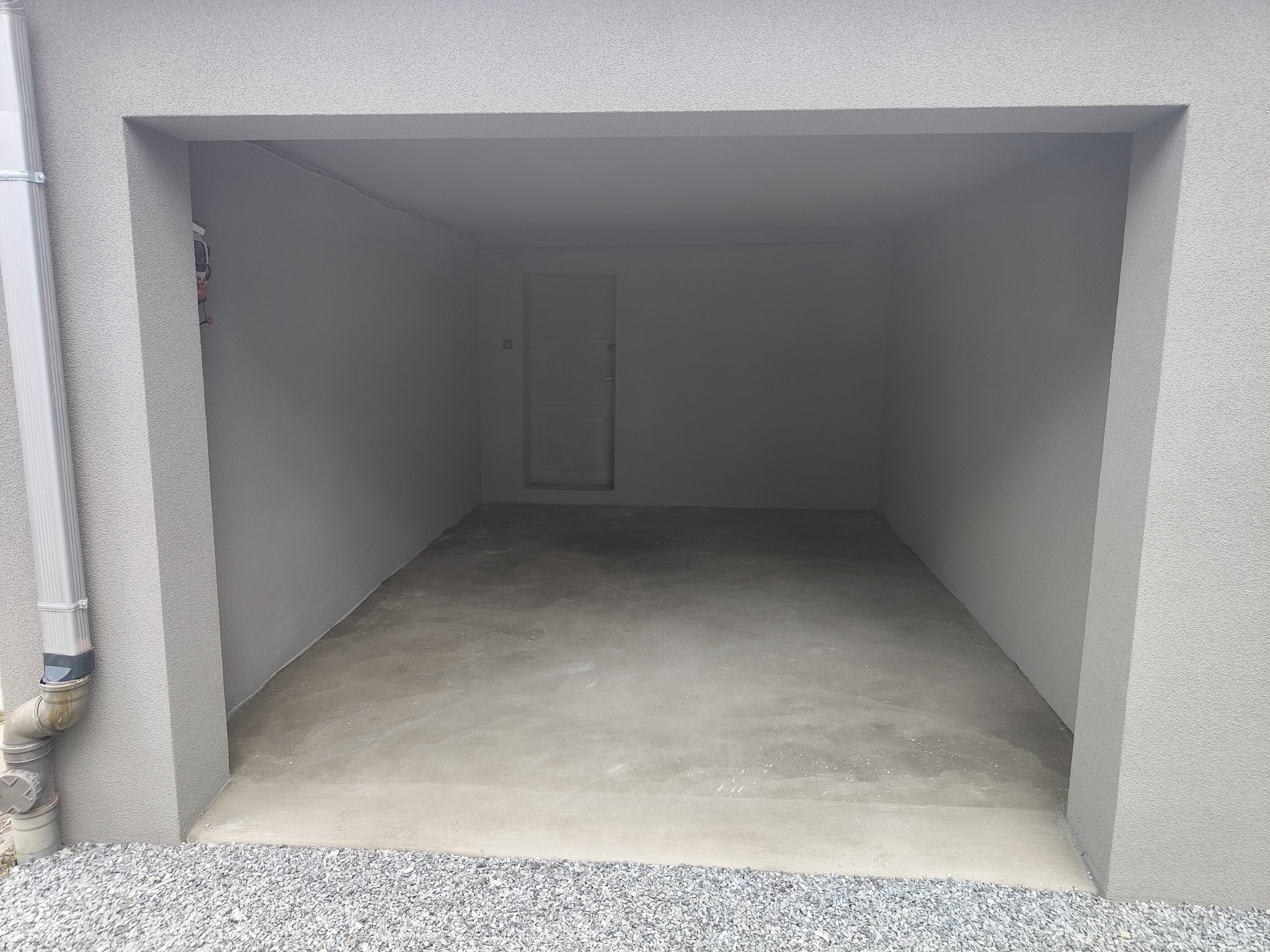 Wnętrze garażu z szarą, teksturowaną ścianą i świeżo wylaną, gładką posadzką. Na zewnątrz widoczne kamienie i rura spustowa.